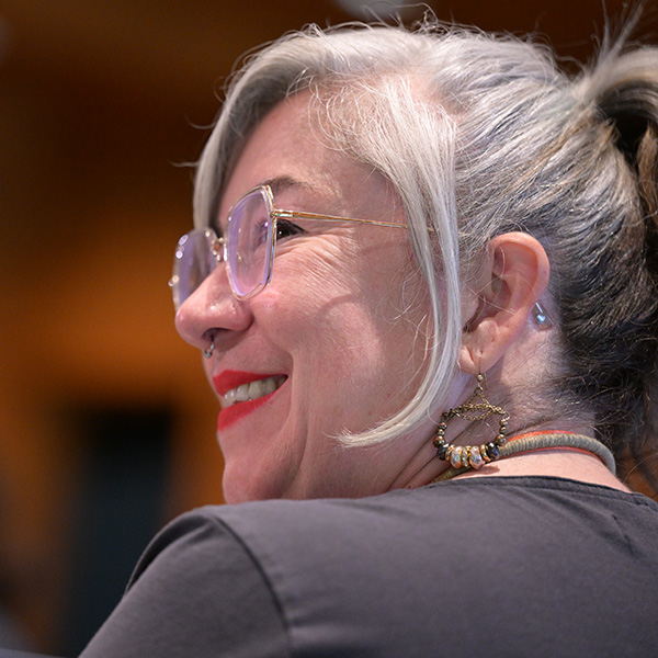 Smiling woman with gray hair tied back, wearing glasses, hoop earrings with beads, and red lipstick, viewed from the side in an indoor setting.