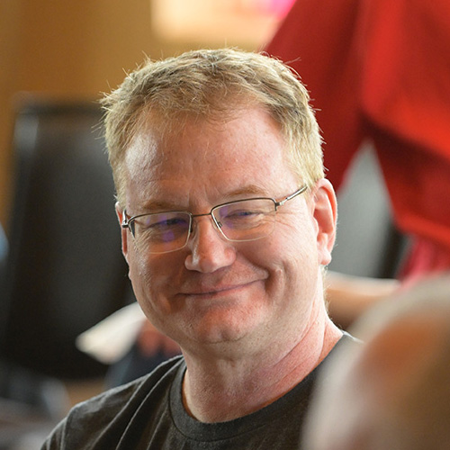 Smiling man with short blond hair and glasses wearing a dark t-shirt, seated indoors with other people in the background.
