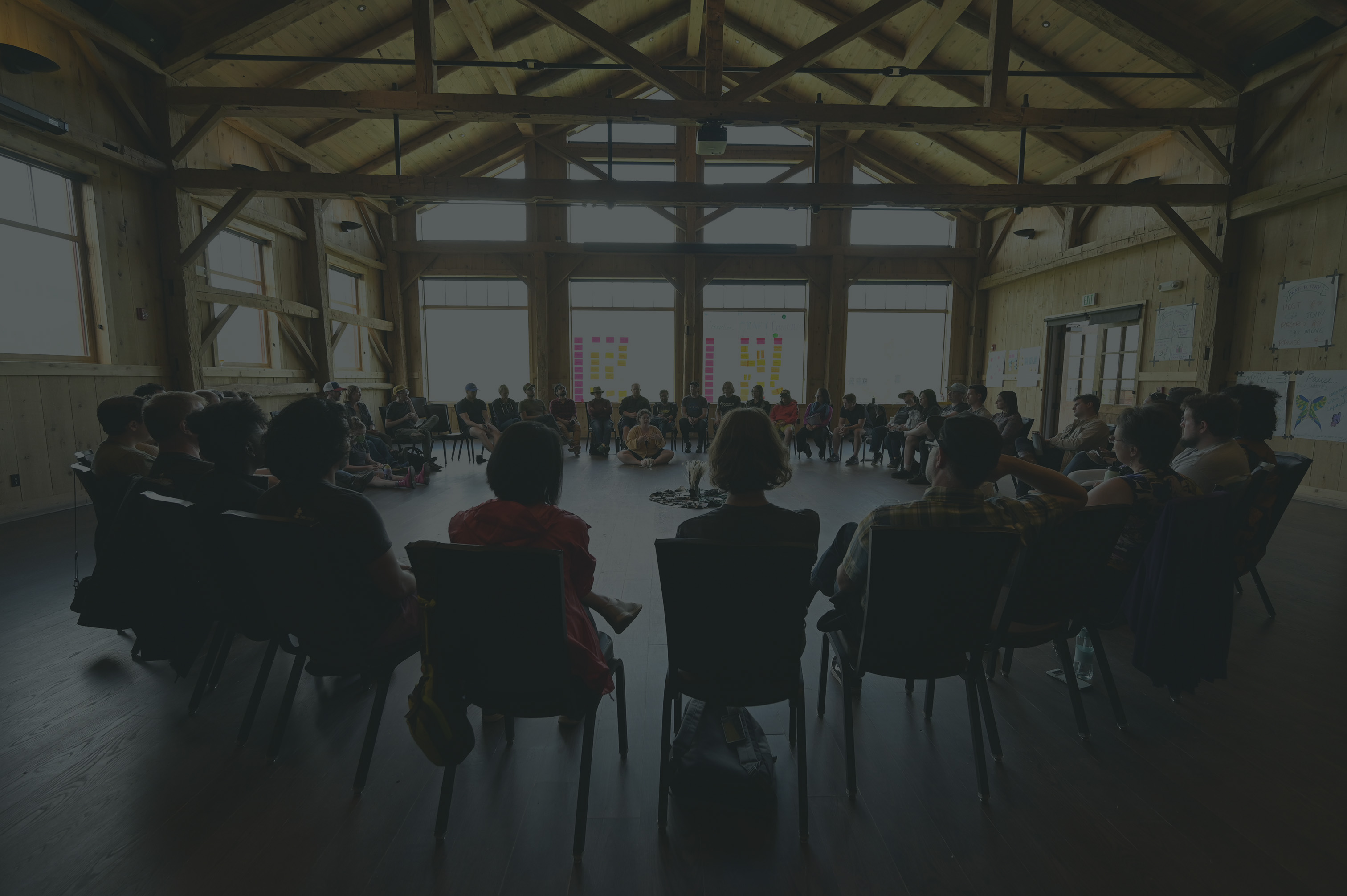A large group of people sits in a circle in a rustic wooden hall with high ceilings and natural light. Colorful sticky notes line the windows, and posters decorate the walls, suggesting a collaborative workshop or group discussion.