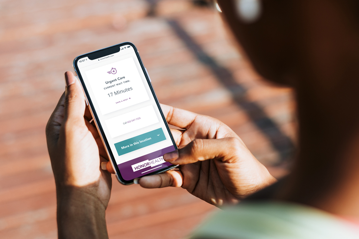 A person is holding a smartphone with a healthcare app open on the screen. The app displays 'Urgent Care Current Wait Times: 17 Minutes' with a call button and additional options for more information on the location, indicated by the name 'HONORHEALTH' at the bottom.
