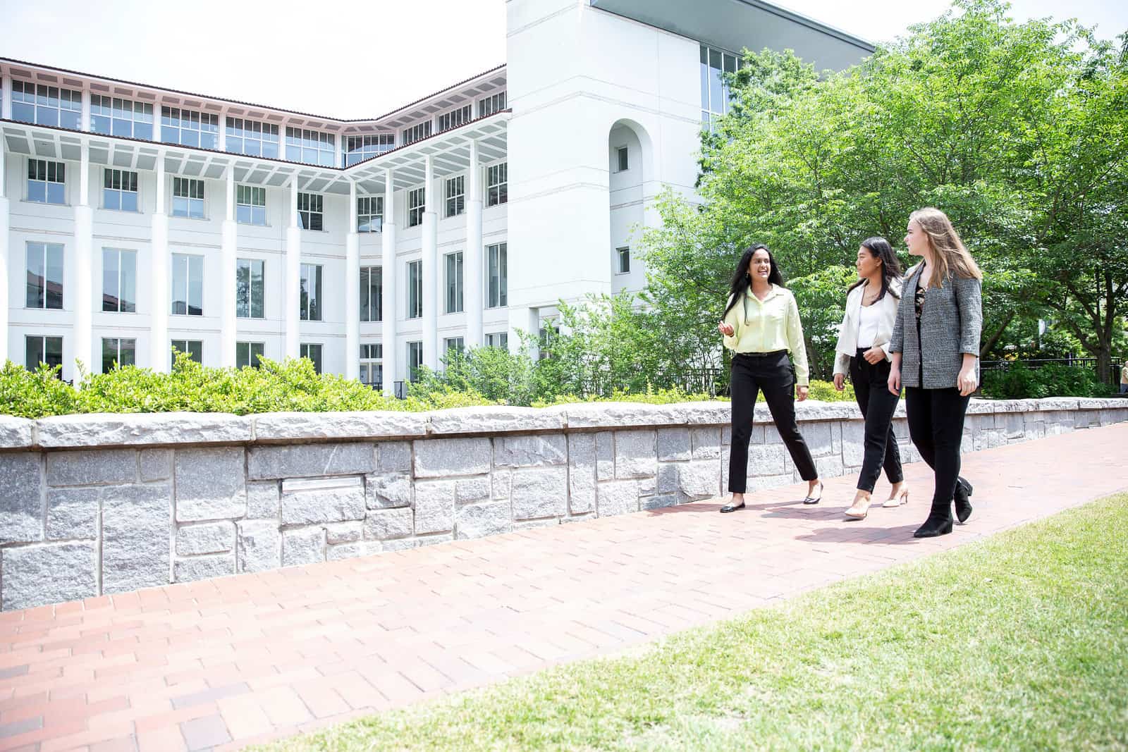 Three women in business attire walking together on a brick pathway on the Goizueta campus outside a large white office building with tall windows and greenery in the background.