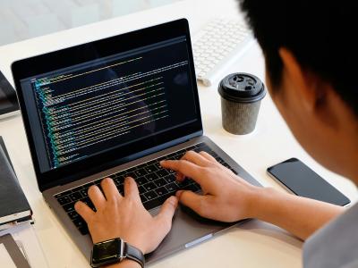 Over-the-shoulder view of a person typing on a laptop displaying multiple lines of code on a dark background. The workspace is clean and white, featuring a smartphone, a cup of coffee, a tablet, and a notebook alongside the computer.
