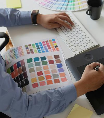 High-angle view of a designer at a white desk using a graphics tablet and stylus while referencing a color swatch sheet. The workspace includes a wireless keyboard, a desktop monitor, a coffee mug, and a set of colorful pastels.