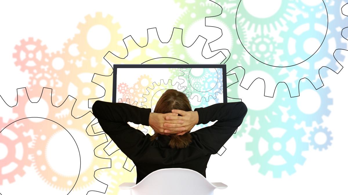 Photo of a person with their hands on the back of their head, looking at a screen with drawings of gears in the background