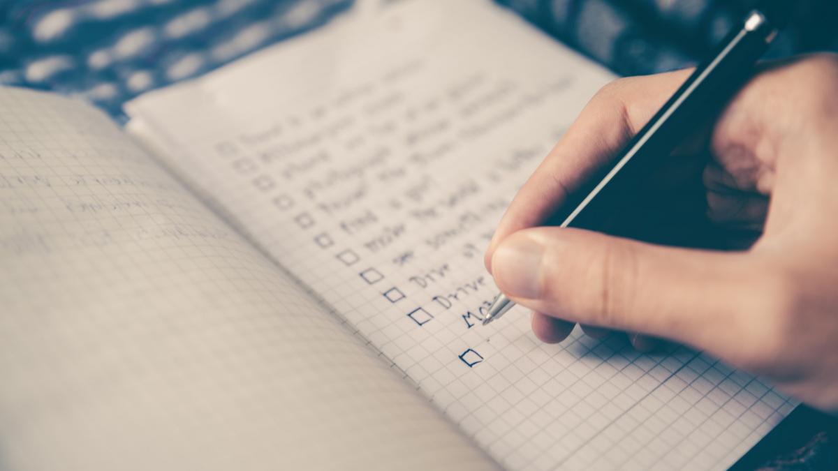 Closeup photo of a person's hand holding a pen and writing a checklist in a notebook