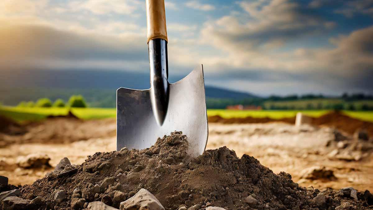 A shovel with a wooden handle and a shiny metal head is plunged into the ground at a construction site. The soil is rocky and uneven, with clumps of dirt and scattered stones around the shovel's blade. In the background, there's a partially overcast sky above a rural landscape with green fields and distant hills.