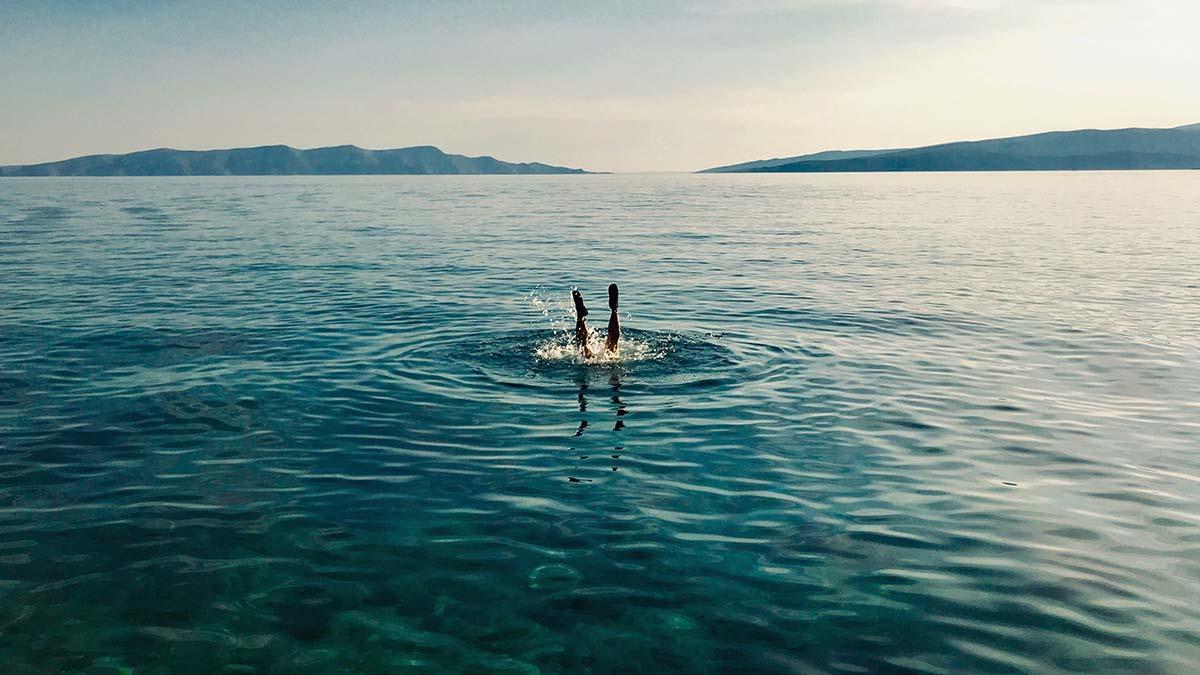 A person's legs and feet stick straight out of the surface of a calm, blue-green ocean. Ripples spread from where their body enters the water, with mountains in the distance.