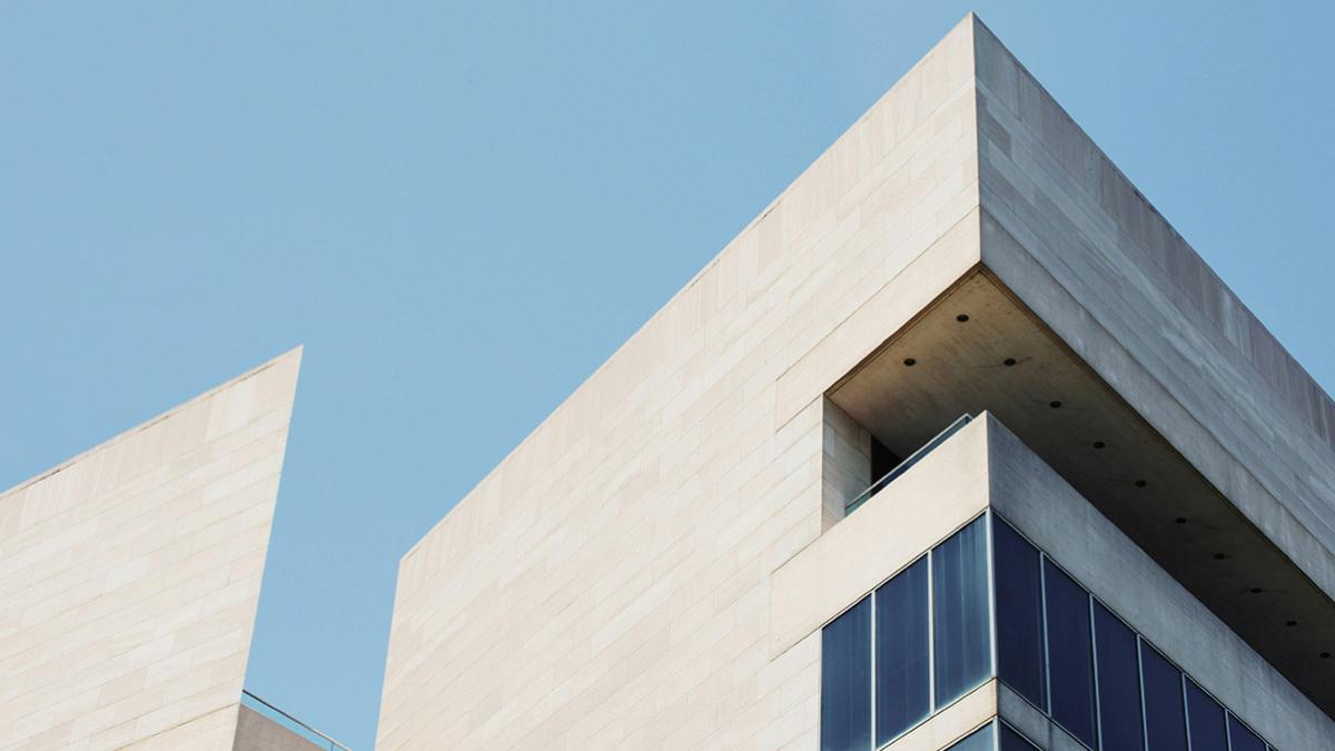 Modern architectural building with clean lines and a minimalist design. The structure features light-colored stone walls and large glass windows, set against a clear blue sky. The design includes sharp angles and an extended balcony area.