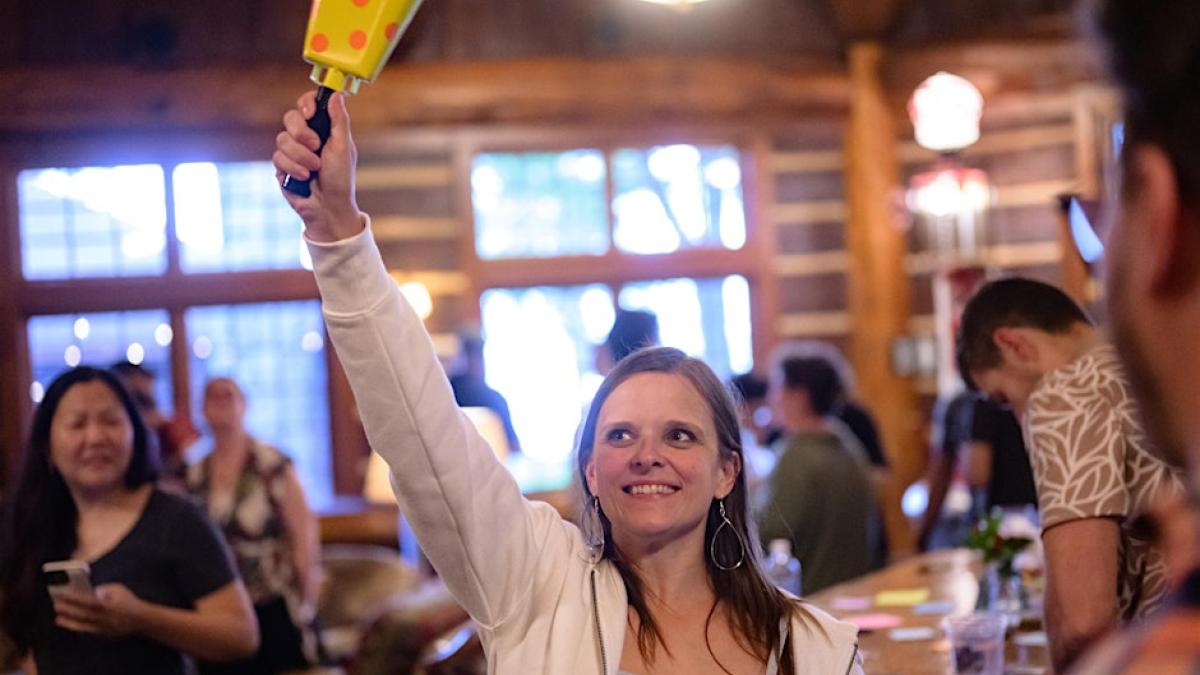 A smiling woman in a white hoodie raises a yellow polka-dotted bell in a rustic lodge. Others are visible in the blurred background.