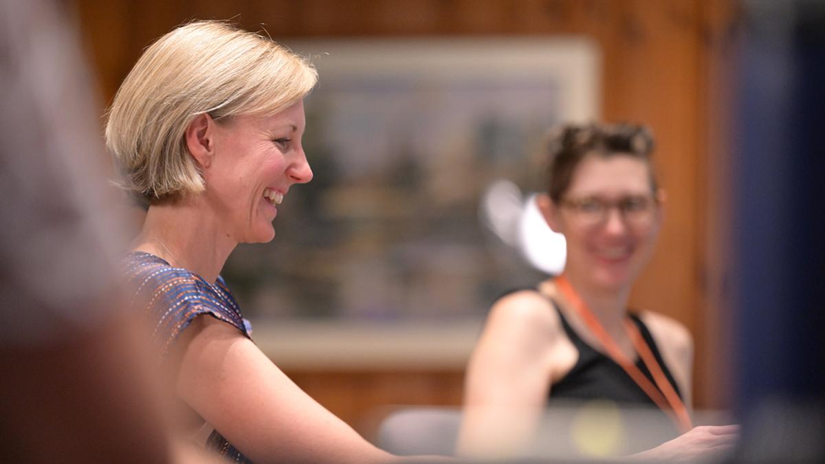 Alt Text for Image:  Two women are seated indoors, smiling and engaged in conversation. The woman in the foreground has short blonde hair and is wearing a colorful patterned top, while the woman in the background, slightly out of focus, has short brown hair, glasses, and a black sleeveless top with an orange lanyard. The setting appears warm and casual, with a wooden wall and framed artwork in the background.
