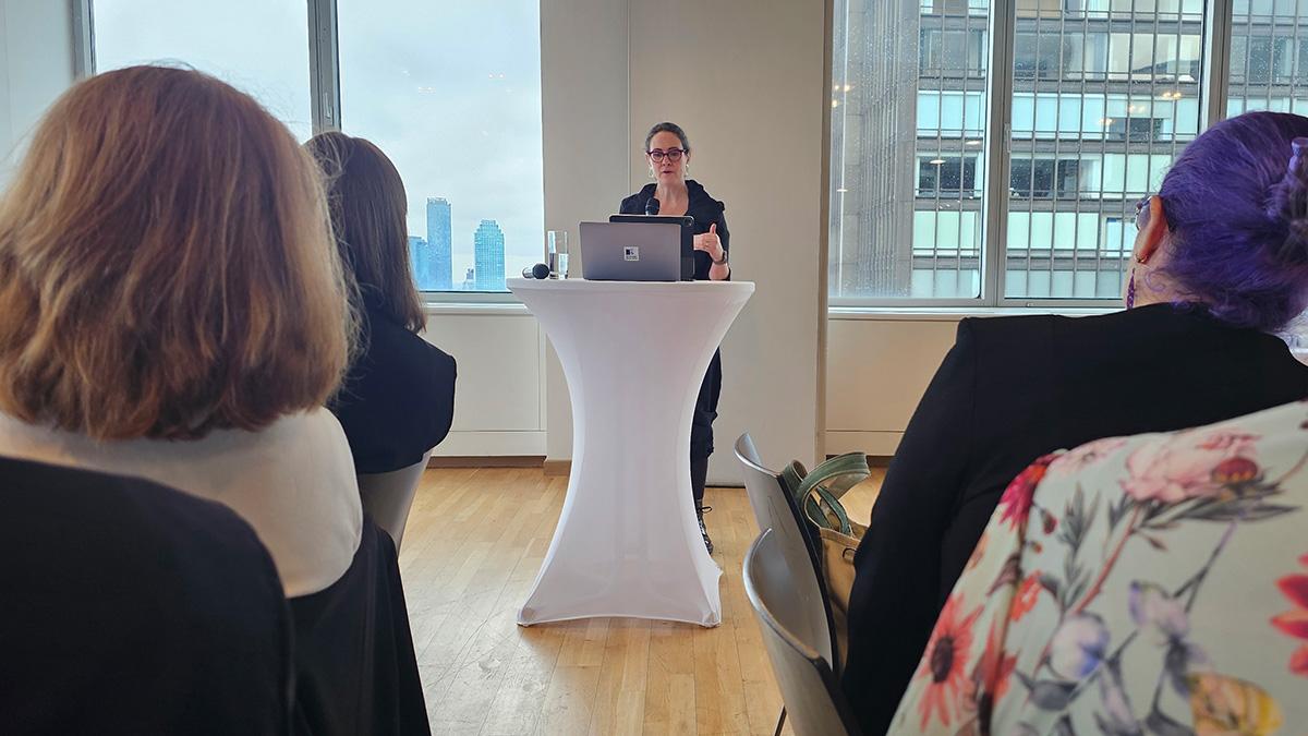Tiffany Farriss speaks at a white podium with a laptop, addressing an audience in a room with large windows overlooking a city skyline.