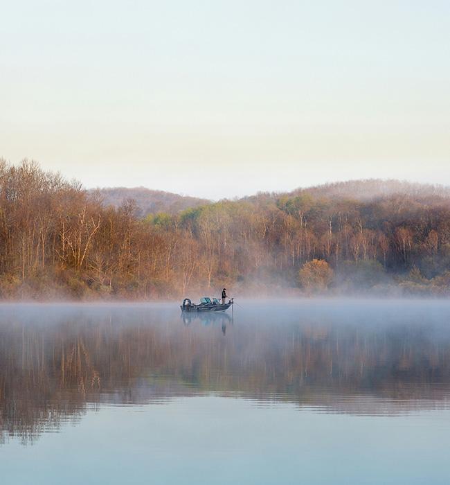 A lone fisherman in a boat on a misty lake, surrounded by bare trees and calm reflections in the early morning light.