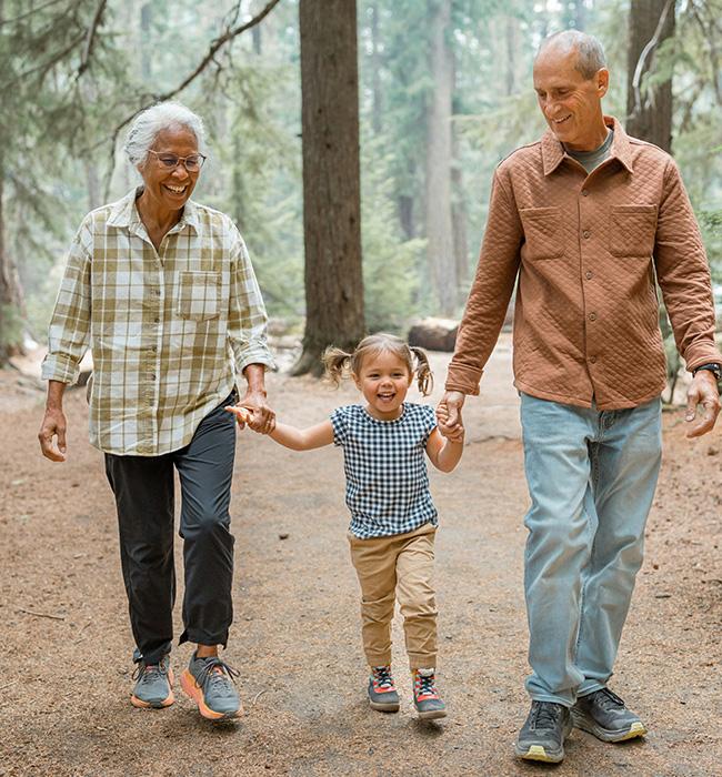 A young child holds hands with two adults, likely grandparents, as they walk along a dirt path in a sun-dappled forest. Tall evergreen trees surround them.