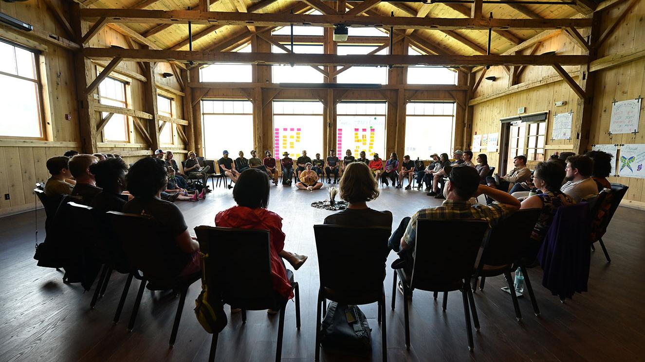 A large group of people sits in a circle in a rustic wooden hall with high ceilings and natural light. Colorful sticky notes line the windows, and posters decorate the walls, suggesting a collaborative workshop or group discussion.
