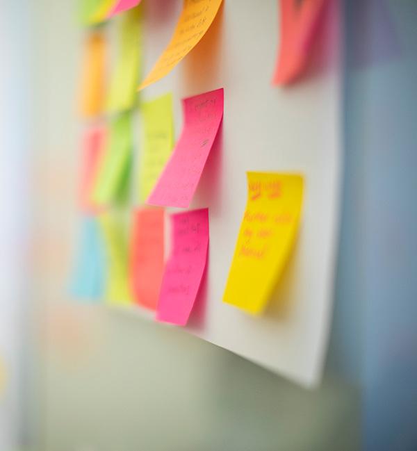 A collection of colorful sticky notes in shades of pink, yellow, orange, and green attached to a white board. The image has a shallow depth of field, with a pink and yellow note in sharp focus while others are blurred, suggesting a creative brainstorming or UX design session.