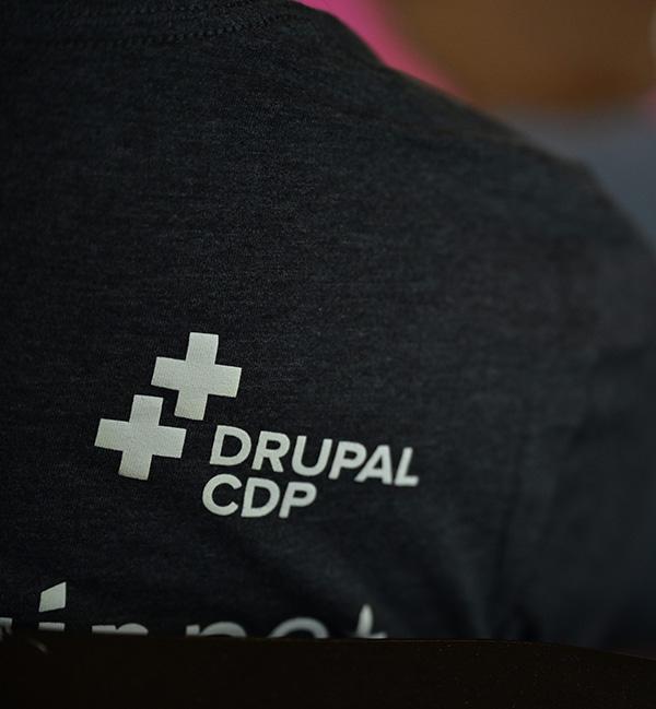 Close-up of the back of a dark gray heathered t-shirt featuring the "DRUPAL CDP" logo in white. The logo includes two overlapping plus signs next to the text. The background is softly blurred.