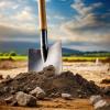  A shovel with a wooden handle and a shiny metal head is plunged into the ground at a construction site. The soil is rocky and uneven, with clumps of dirt and scattered stones around the shovel's blade. In the background, there's a partially overcast sky above a rural landscape with green fields and distant hills.