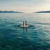 A person's legs and feet stick straight out of the surface of a calm, blue-green ocean.  Ripples spread from where their body enters the water, with mountains in the distance.