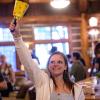 A smiling woman in a white hoodie raises a yellow polka-dotted bell in a rustic lodge. Others are visible in the blurred background.