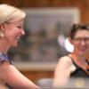 Alt Text for Image:  Two women are seated indoors, smiling and engaged in conversation. The woman in the foreground has short blonde hair and is wearing a colorful patterned top, while the woman in the background, slightly out of focus, has short brown hair, glasses, and a black sleeveless top with an orange lanyard. The setting appears warm and casual, with a wooden wall and framed artwork in the background.