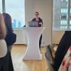 Tiffany Farriss speaks at a white podium with a laptop, addressing an audience in a room with large windows overlooking a city skyline.