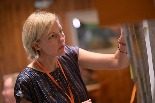Woman with short blonde hair wearing a striped blouse and an orange lanyard, looking intently at a wall with papers or sticky notes, as if reviewing or organizing information.